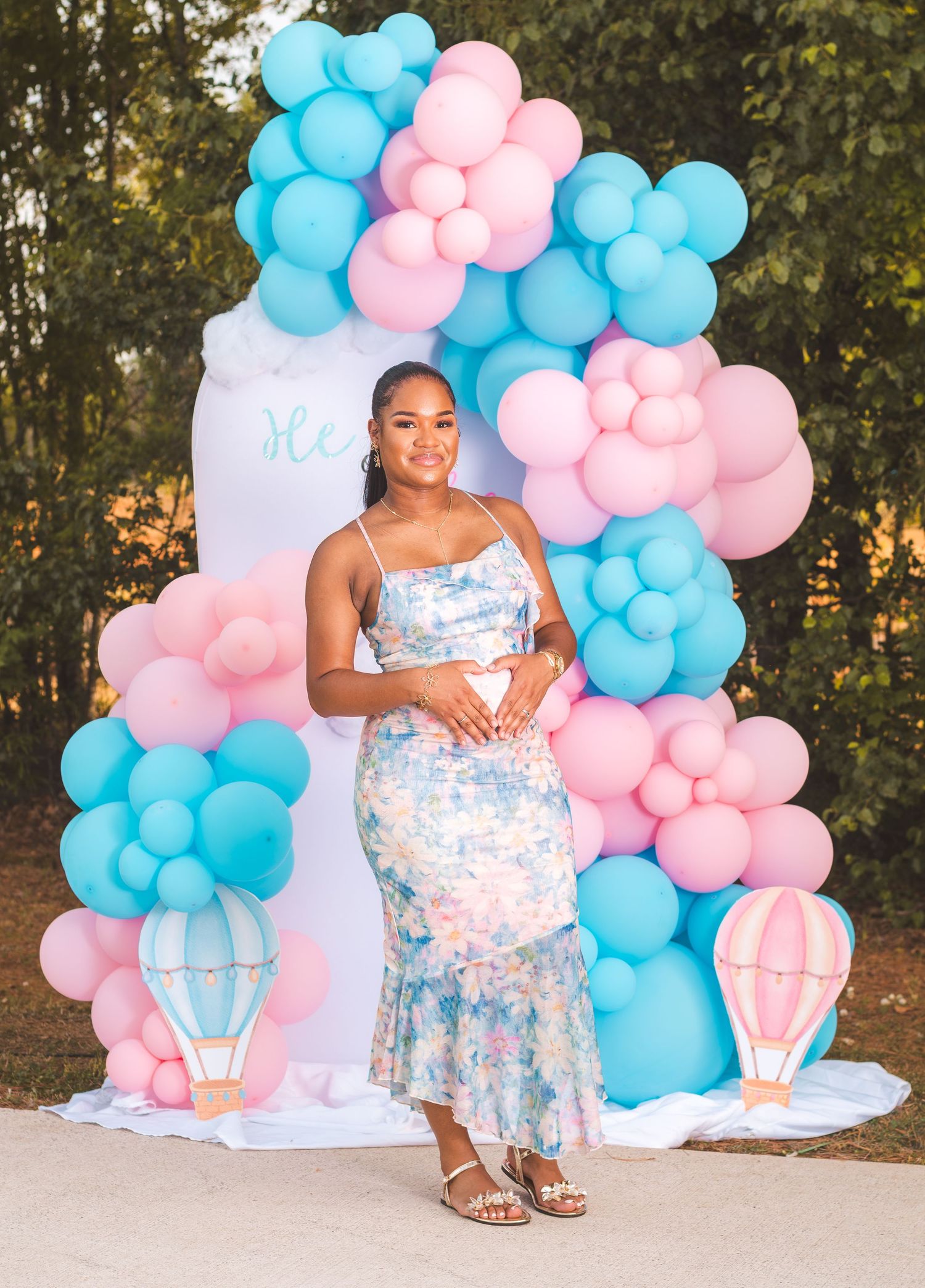 Pregnant woman in floral dress poses at gender reveal party with pink and blue balloon arch backdrop.