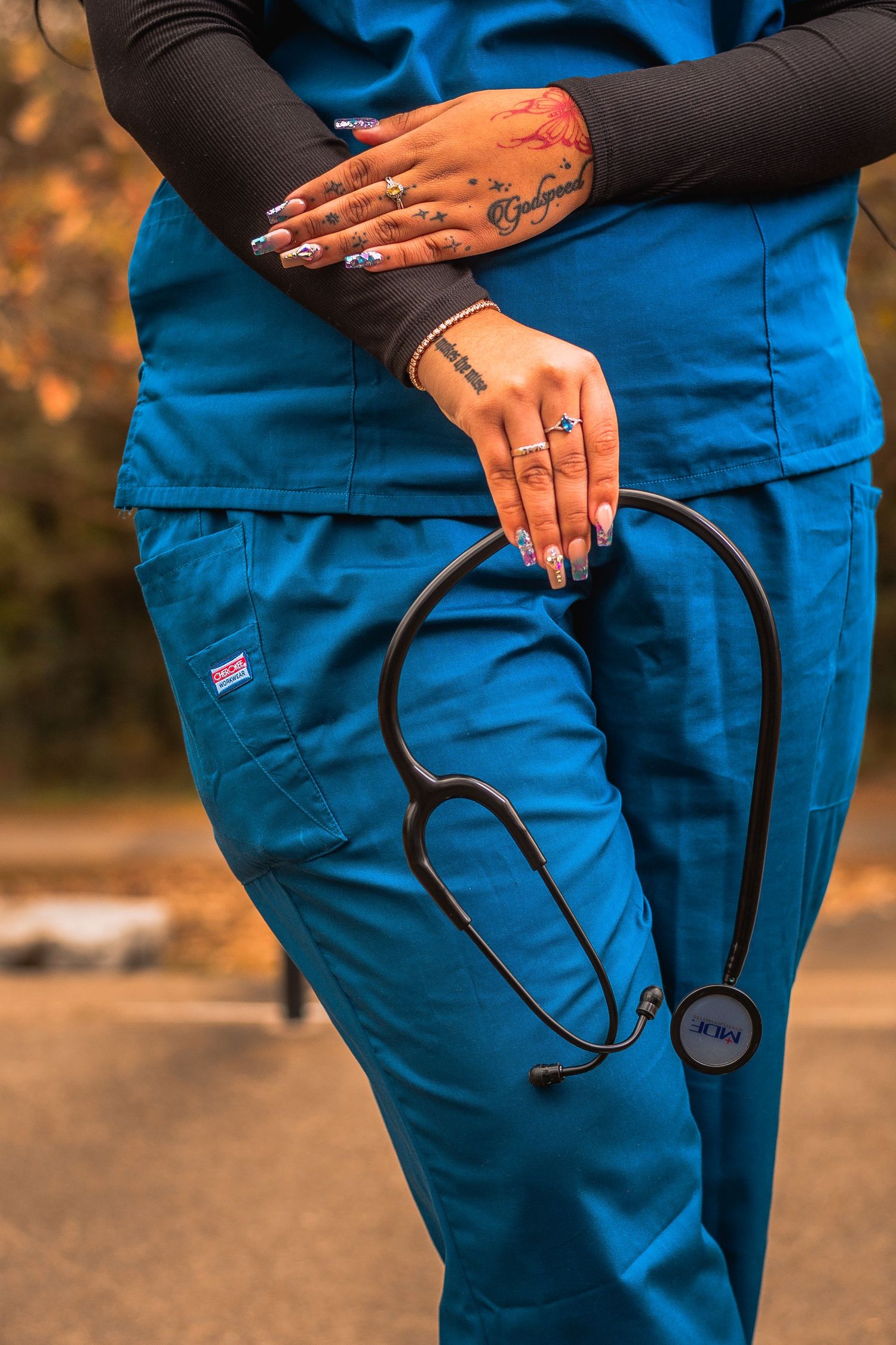 Healthcare worker in blue scrubs holding a black stethoscope, with tattooed hands and colorful nails.