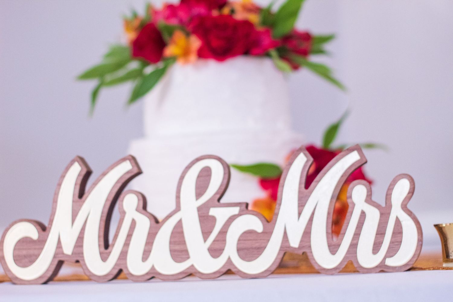 White wooden 'Mr & Mrs' wedding sign decorated with vibrant red and pink flowers on top.