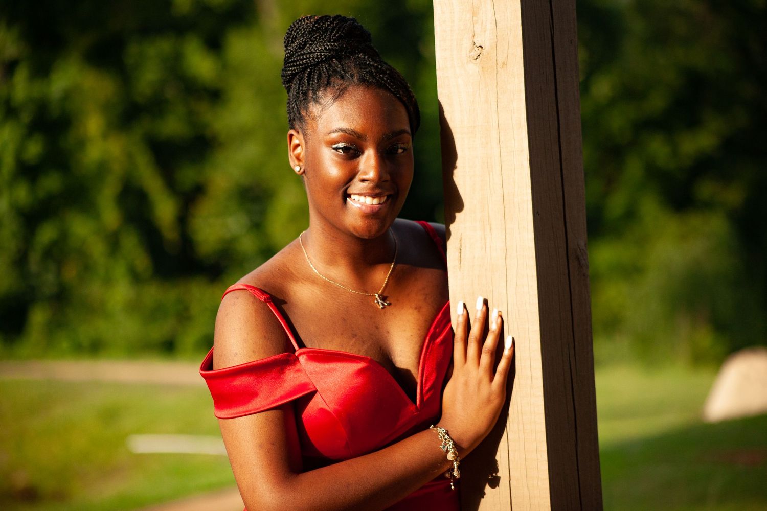 Woman in red off-shoulder dress poses elegantly by wooden post in sunlit garden.