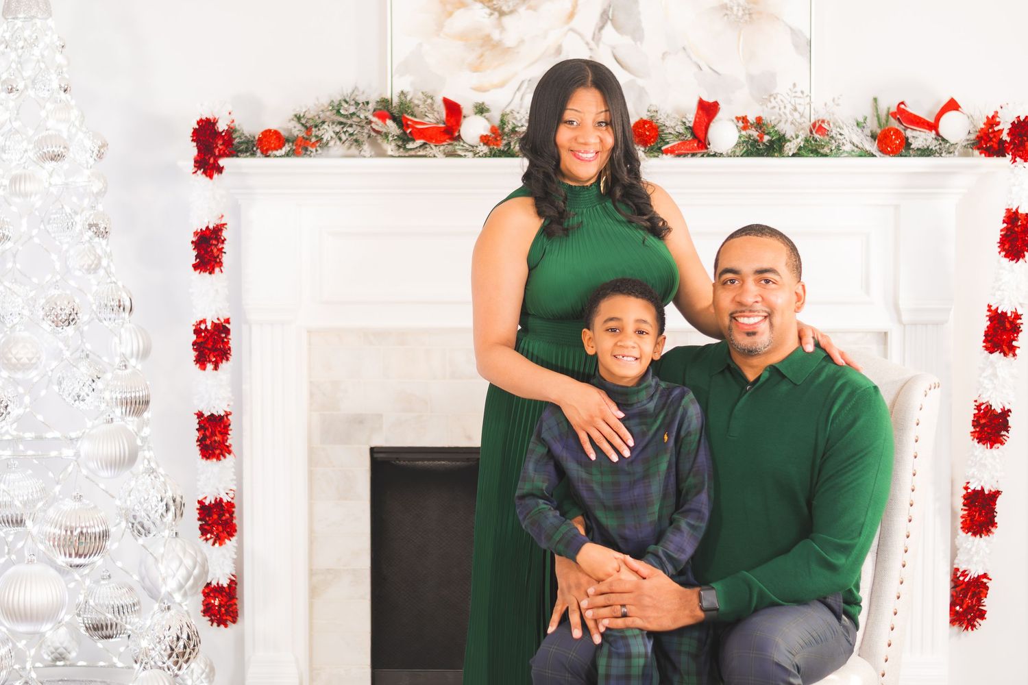Family of three posing for a festive Christmas portrait in front of a decorated white fireplace.