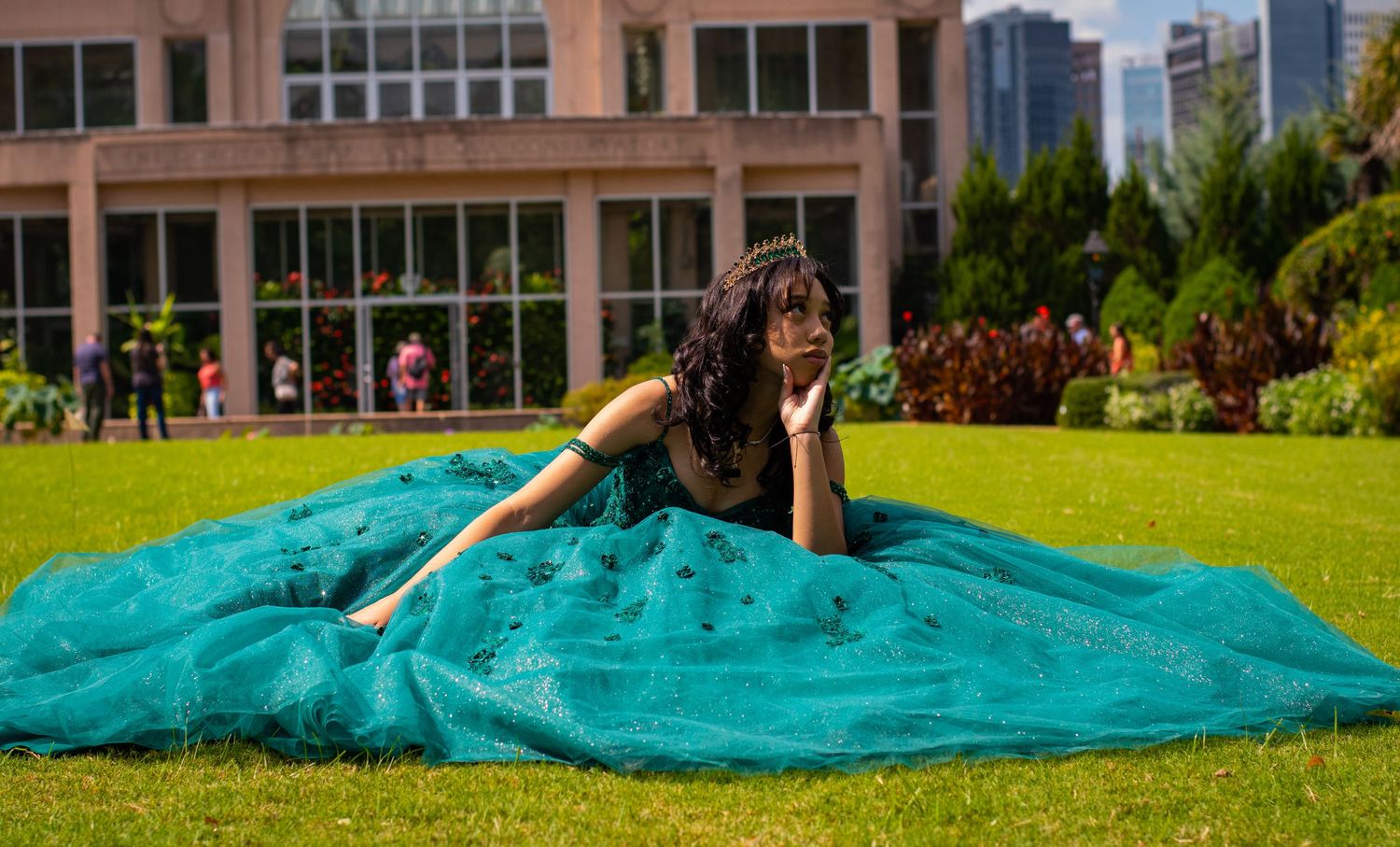 A person in a teal ballgown lies gracefully on lush green grass with city buildings in the background.