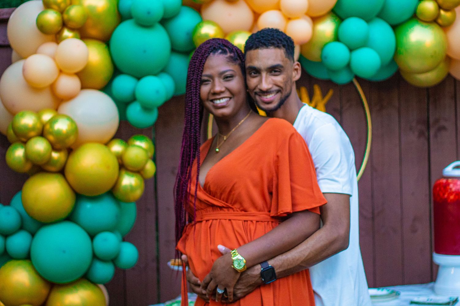 Happy couple at baby shower celebration with festive balloon arch in orange, gold and teal.