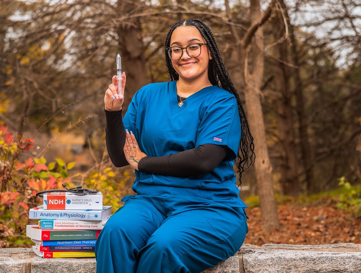 Nursing student in blue scrubs holding syringe, seated with medical textbooks outdoors in autumn setting.
