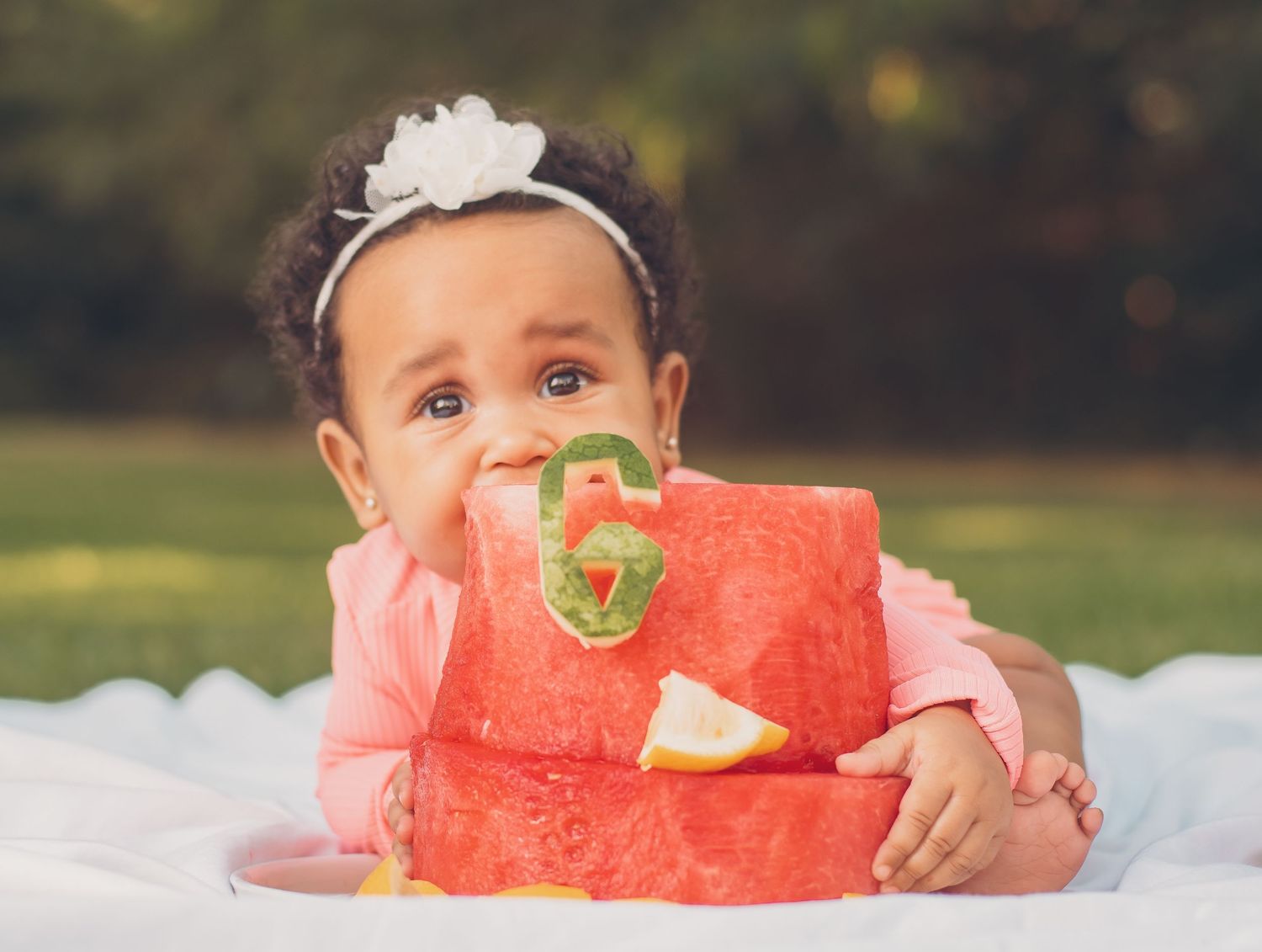 Baby girl in pink outfit biting into a watermelon cake with number 6, celebrating her 6-month milestone outdoors.