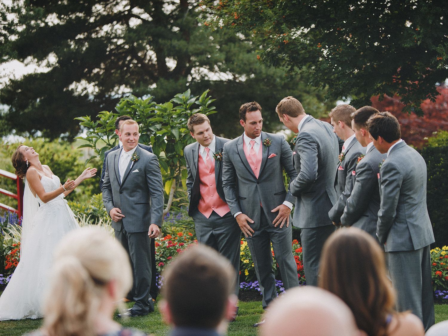 Wedding party members share laughter during an outdoor garden ceremony in grey suits and formal attire.