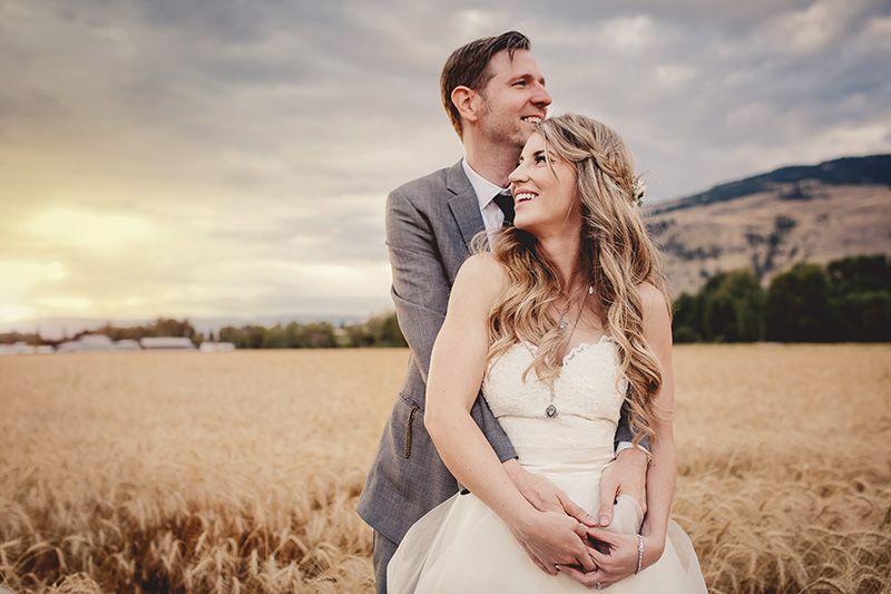 A romantic wedding couple embraces in a golden wheat field at sunset with mountains in the background.