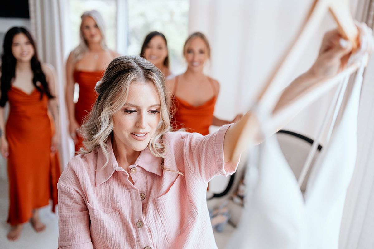 A group of people wearing orange dresses watching a bride in a pink robe as she inspects her wedding gown.