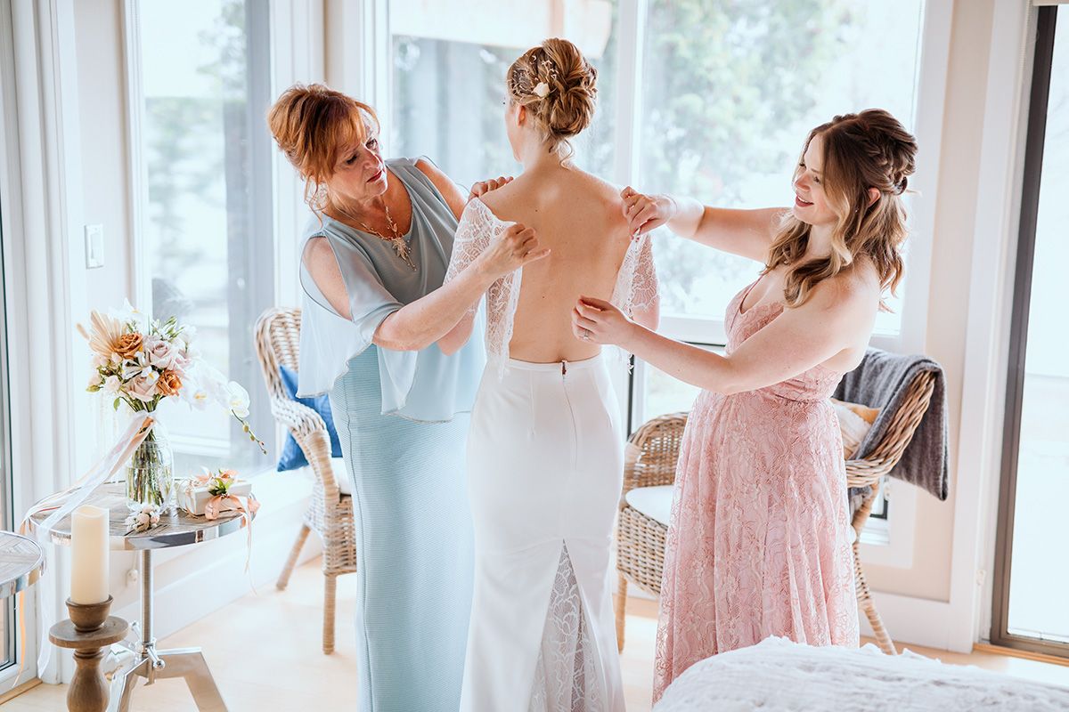 Bridesmaids in pastel blue and pink dresses help each other get ready in a bright sunlit room.