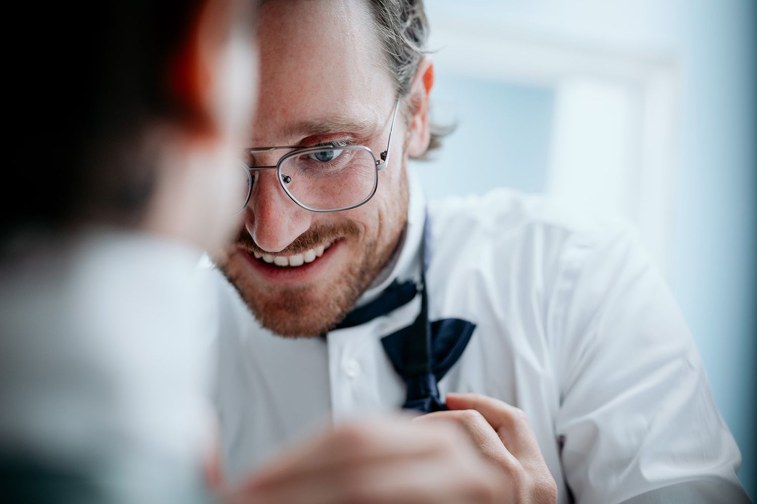 A groom in a white shirt and glasses smiling and laughing while attempting to tie his best man's bow-tie.