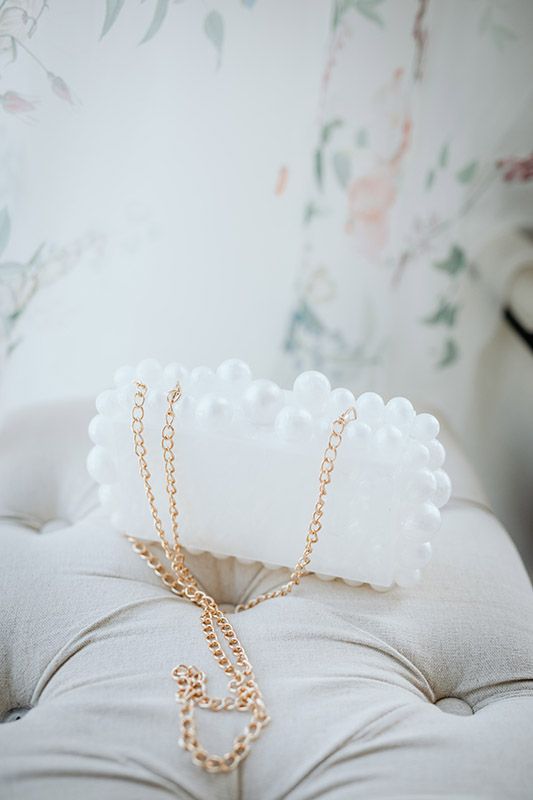 Delicate gold chain laying on soft white clutch hand bag with floral pattern in background.