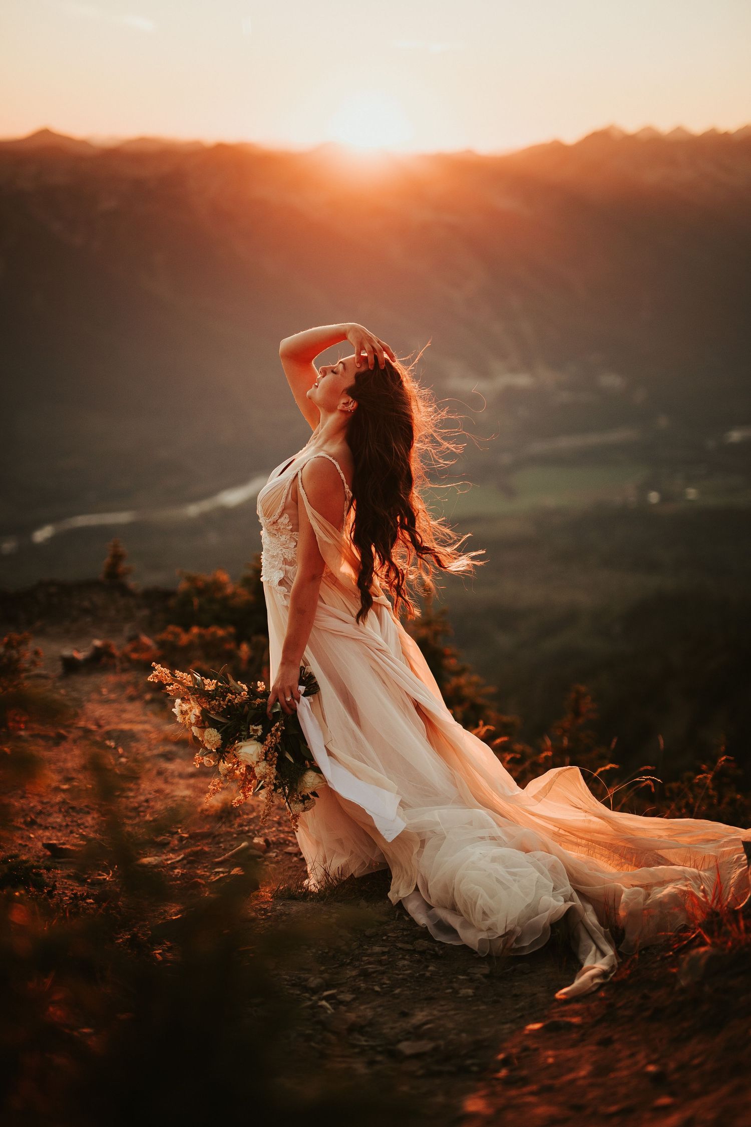 A bride in a flowing white dress sits on a mountaintop at sunset as wind blows through their hair.