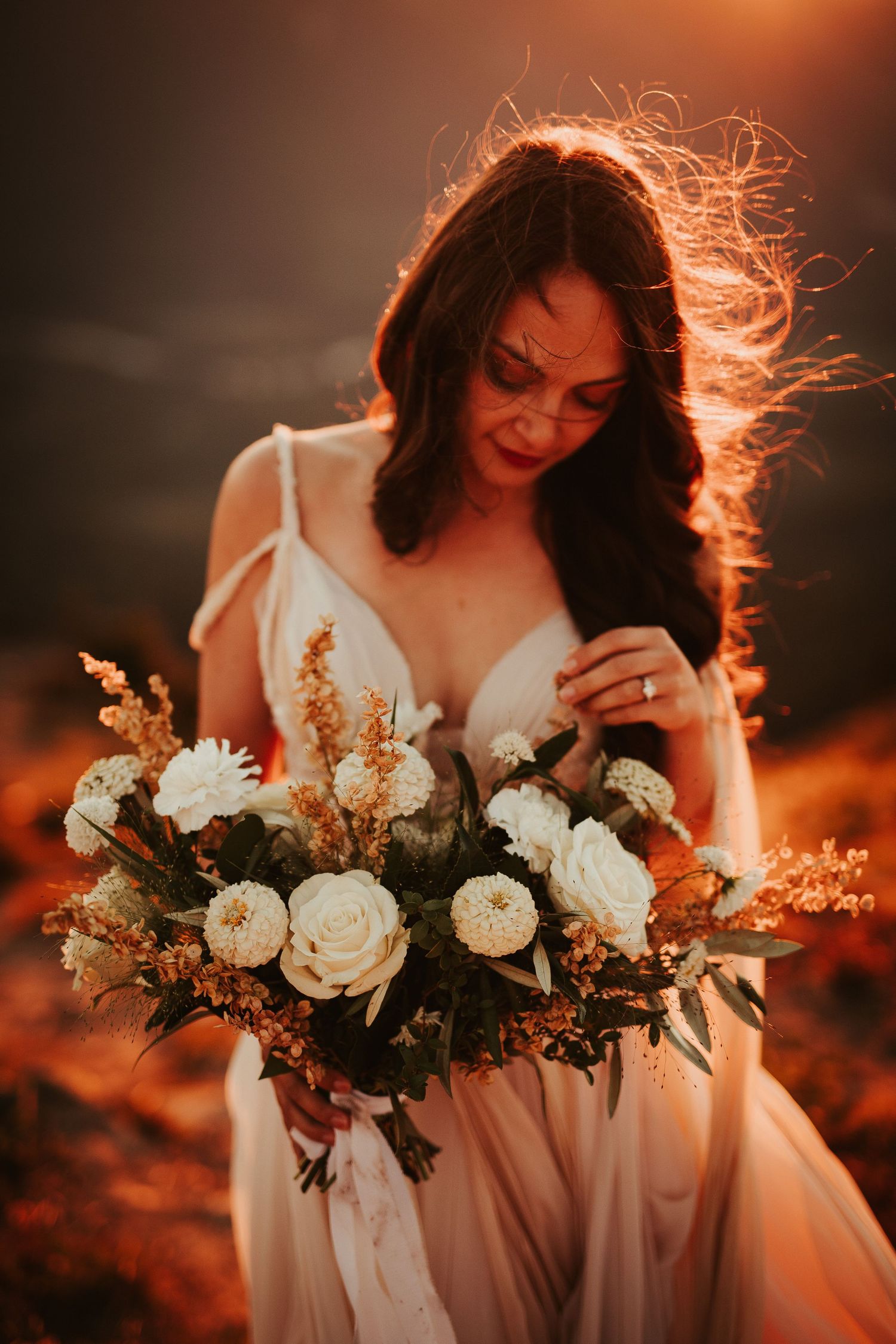 A bride in a white dress holds a romantic bouquet of white roses against a dramatic sunset-lit backdrop.