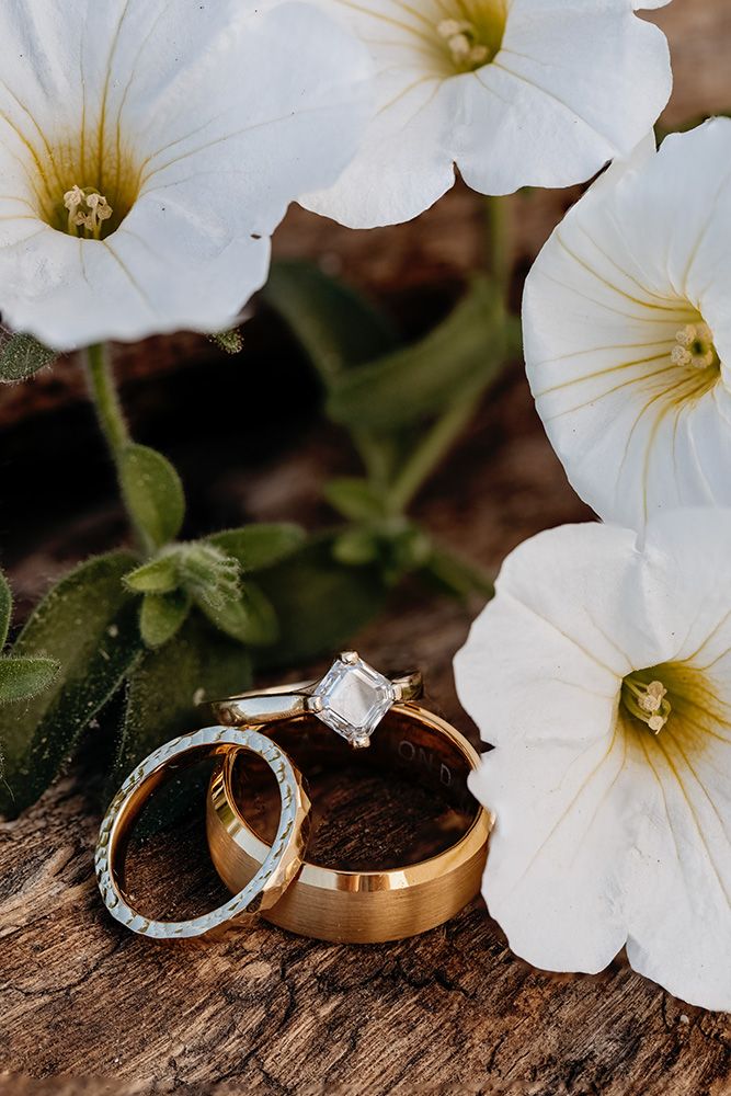 Wedding rings with white petunia flowers arranged on rustic wooden surface in natural lighting.