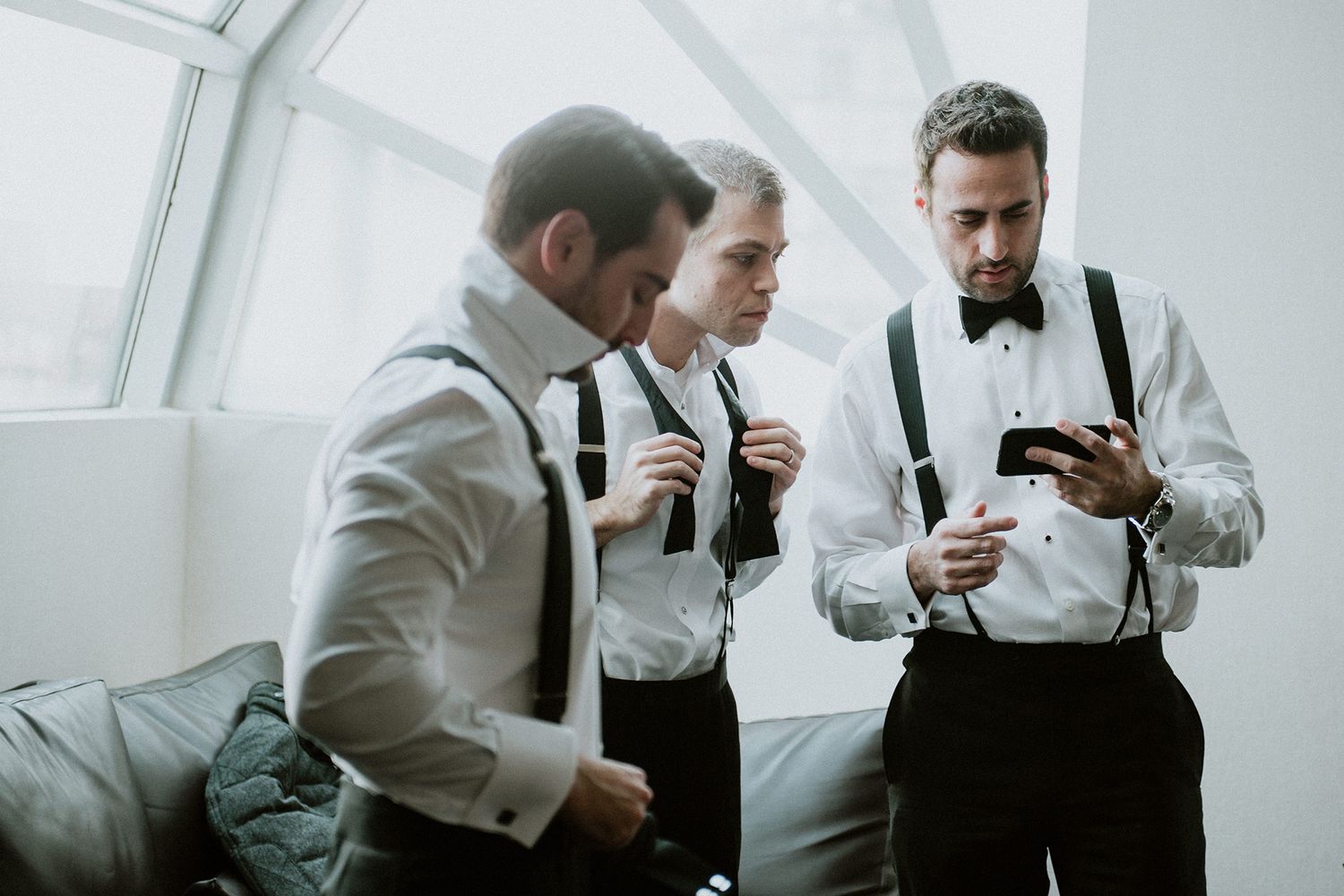 Groomsmen in white shirts and suspenders help each other get ready for a wedding ceremony.