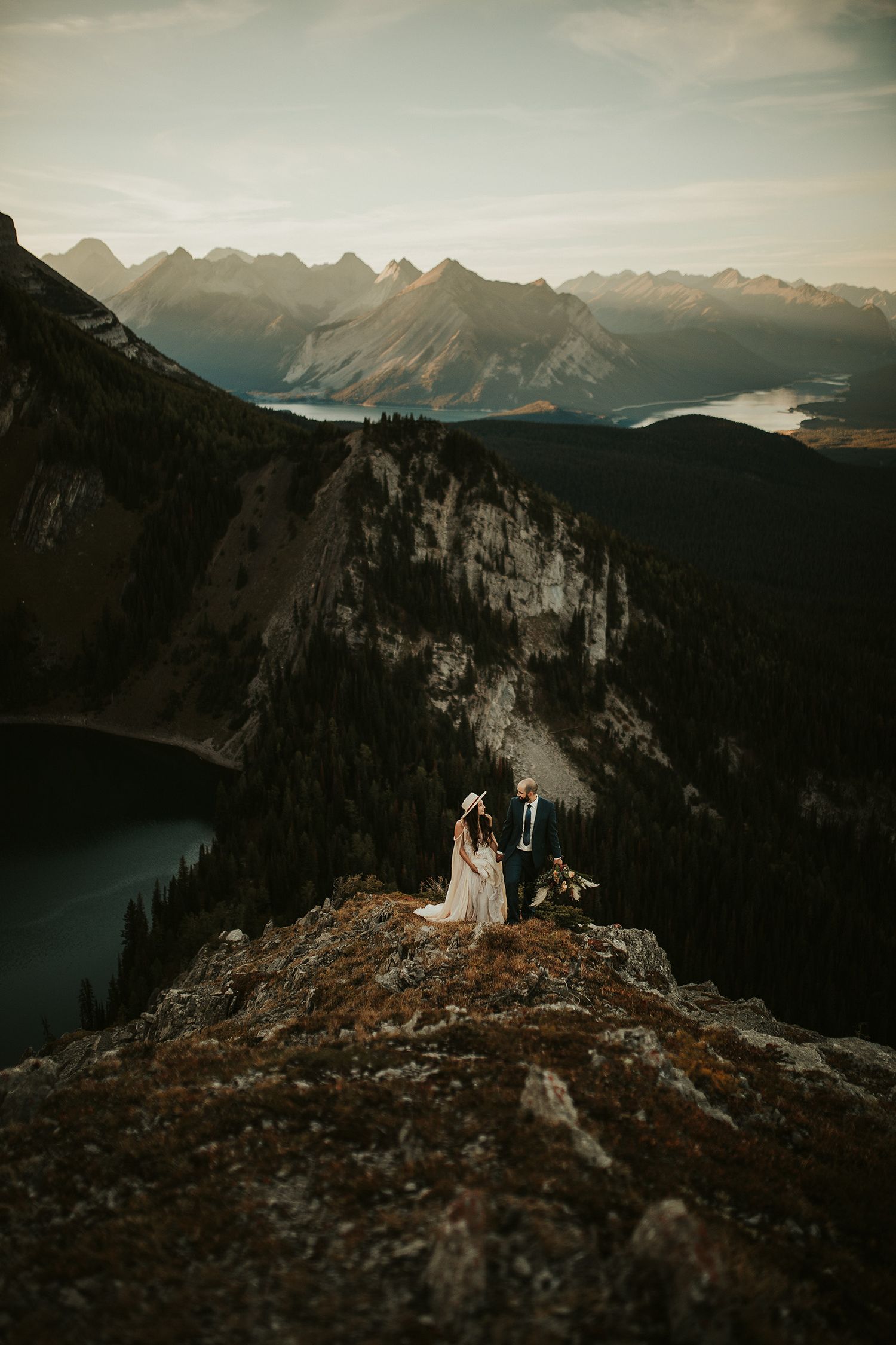 A wedding couple stand on a rocky mountain summit overlooking dramatic alpine peaks and lakes at sunset.