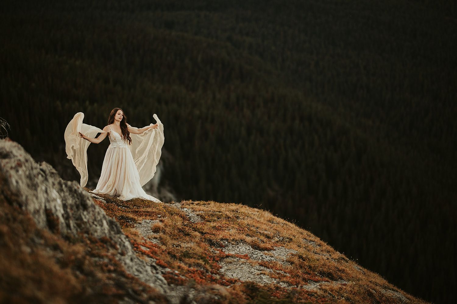A bride poses on the edge of a cliff as wind blows her wedding dress reminiscent of the sculpture, the Winged Victory of Samothrace.