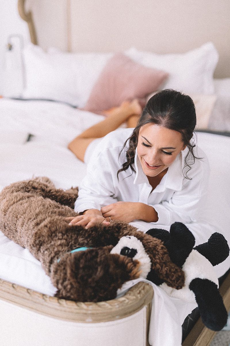 Woman in white pajamas on bed pets her brown poodle dog as it clutches a stuffed toy.