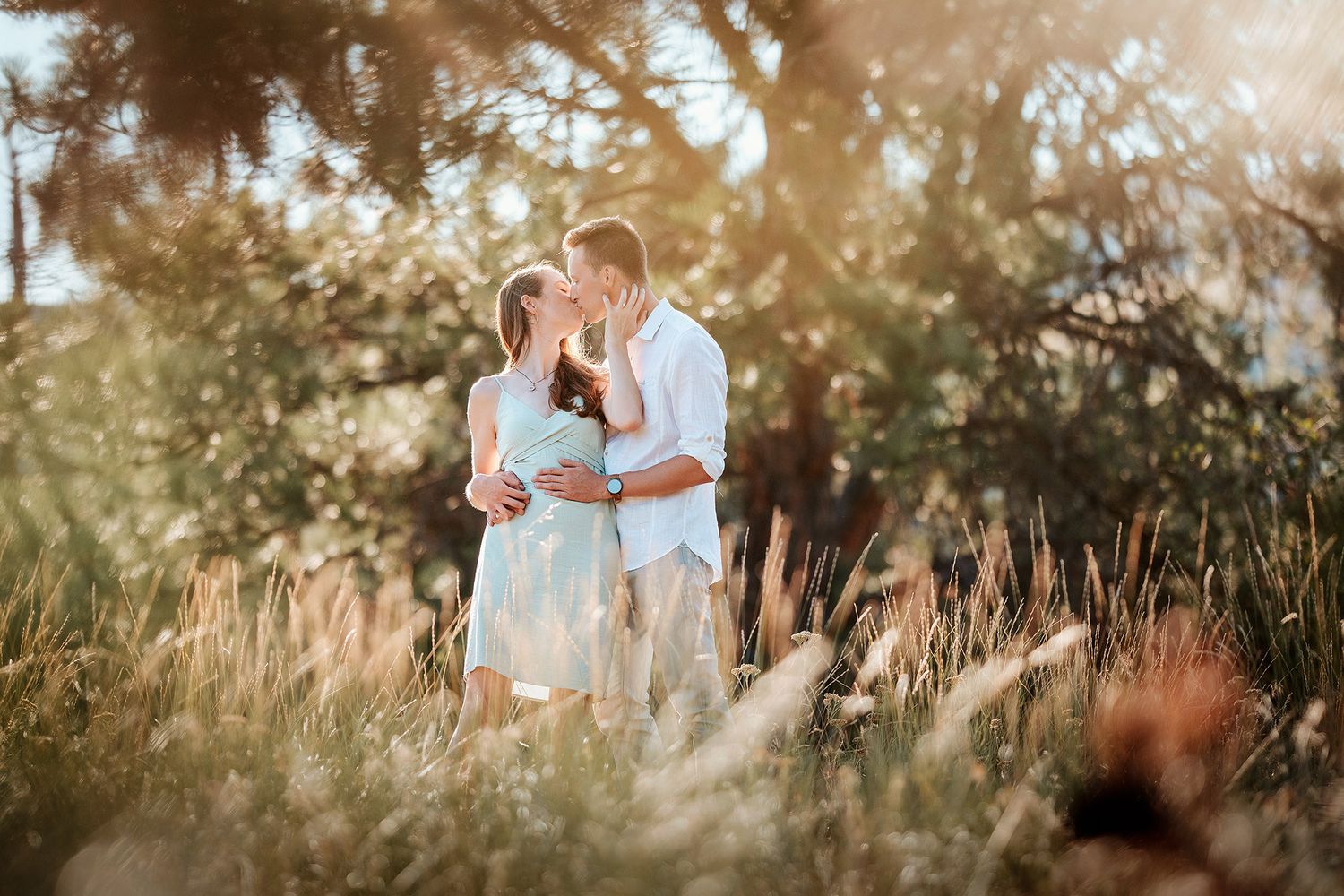 Romantic couple embracing in a sunlit field surrounded by golden grass and trees during a warm summer day.