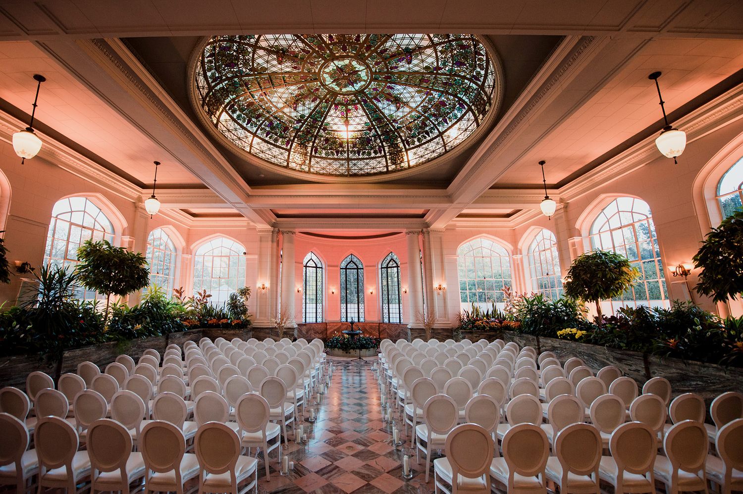 Elegant ballroom with crystal chandeliers, arched windows, and rows of white chairs set up for a formal event.