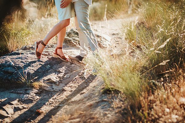 Couple standing close together on a rocky trail surrounded by dry grass, wearing casual summer footwear.