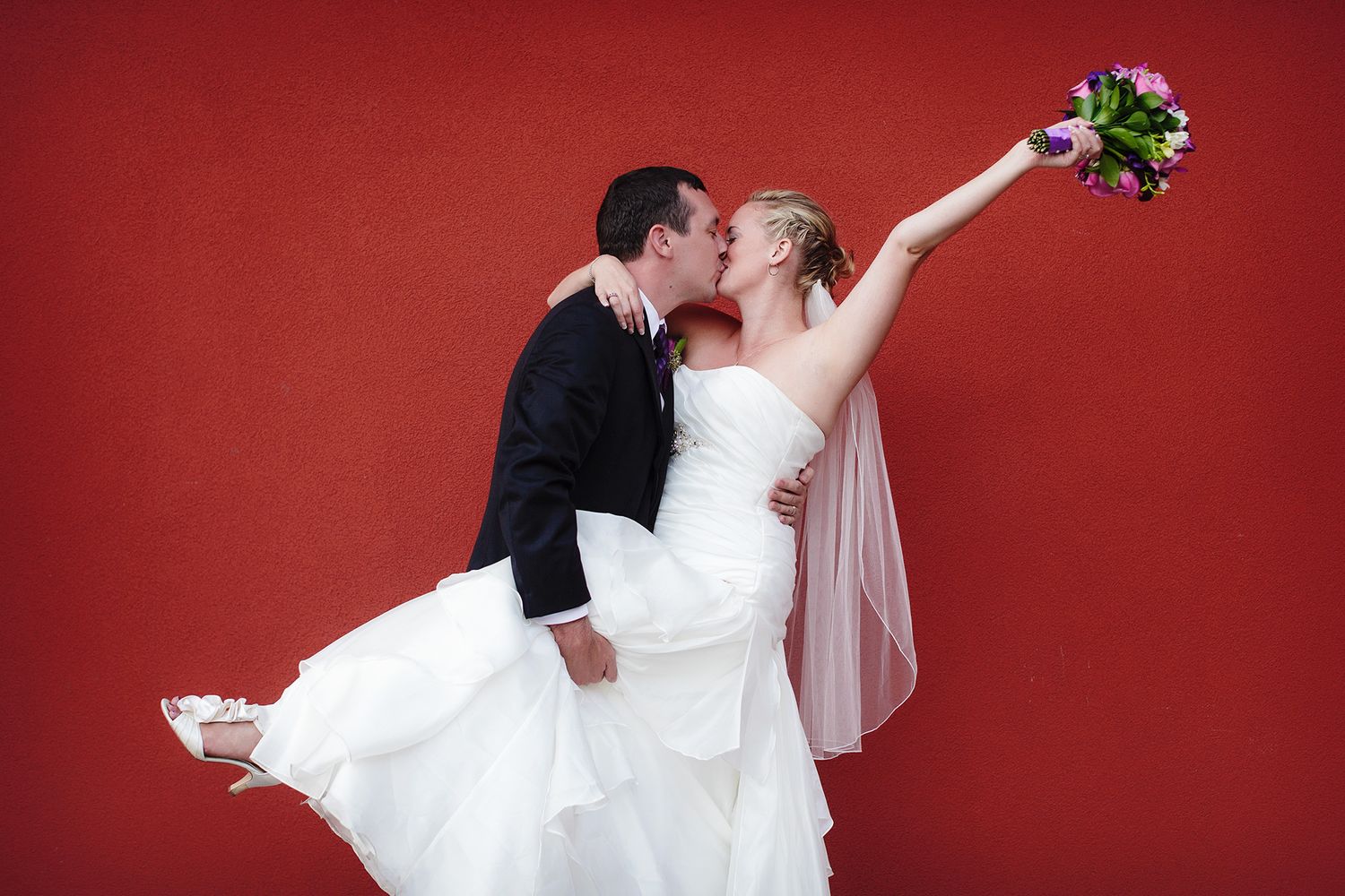 A romantic wedding couple shares a tender kiss against a bold red backdrop.