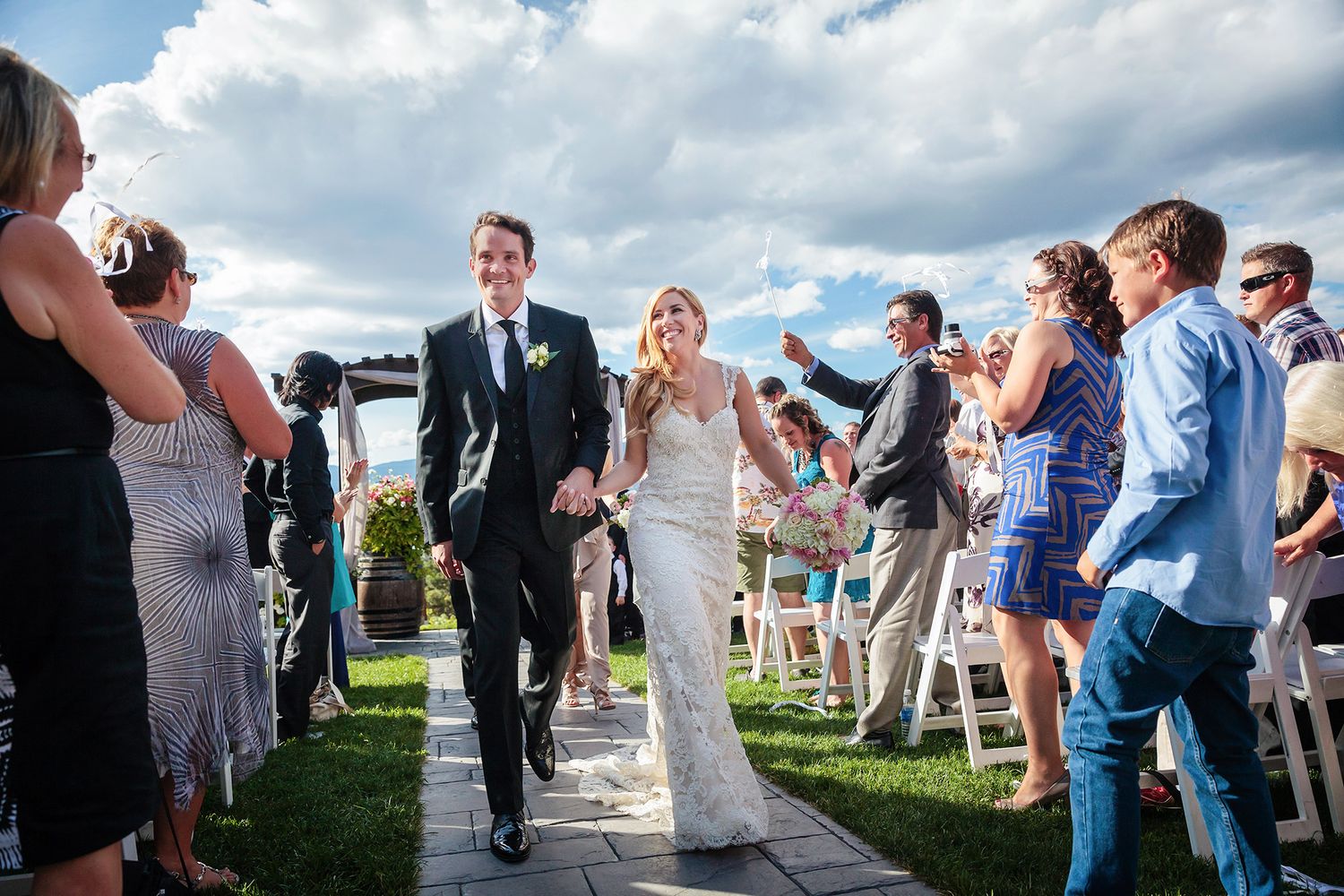 Wedding guests celebrate outdoors under cloudy skies at a scenic vineyard venue during a summer wedding ceremony.