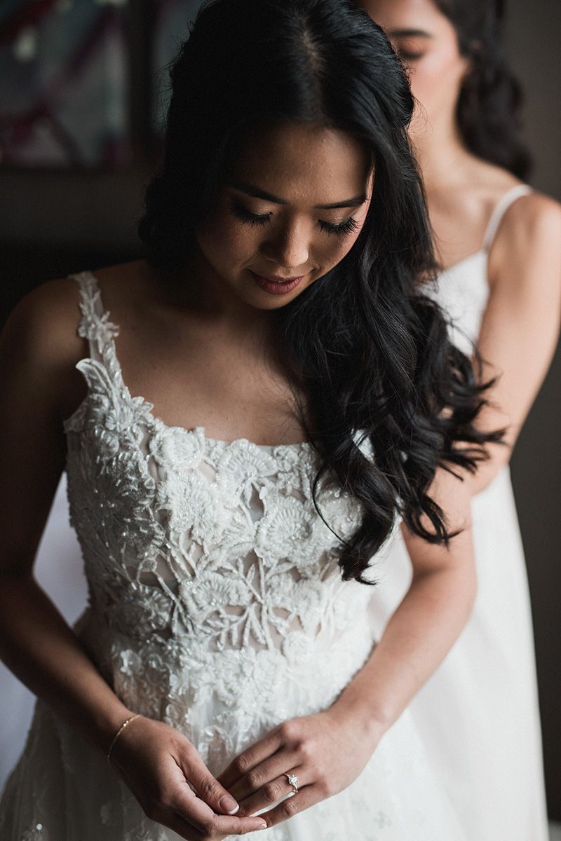 A bride in a delicate white lace wedding gown with floral appliques looks down thoughtfully in soft lighting.