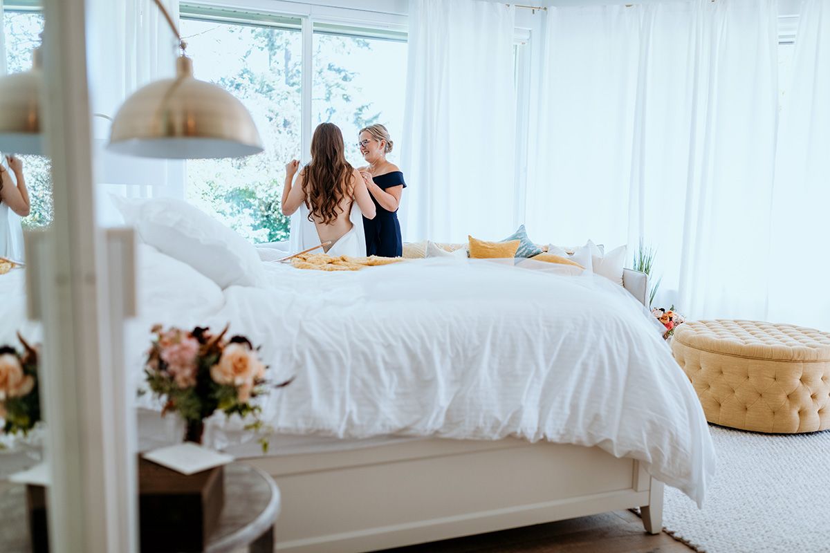 In a bright white bedroom with a plush bed, white linens, and natural light streaming through windows, a bride is helped into her dress.