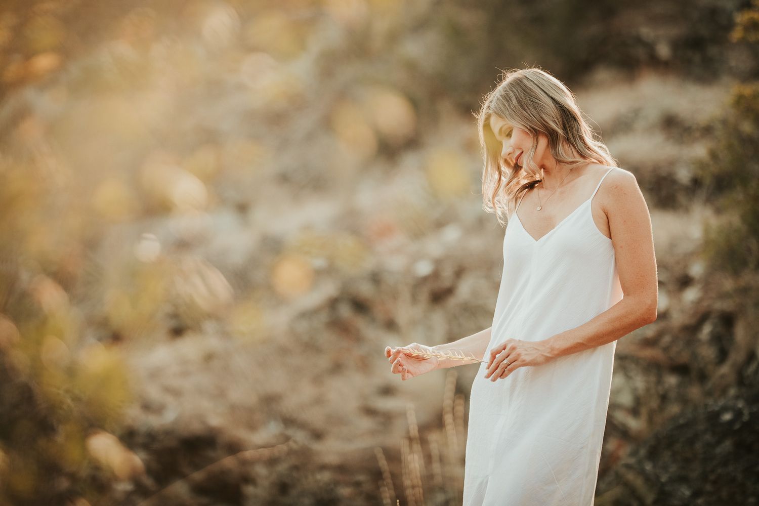 A woman in a flowing white dress stands in a golden field during sunset creating a dreamy ethereal scene.