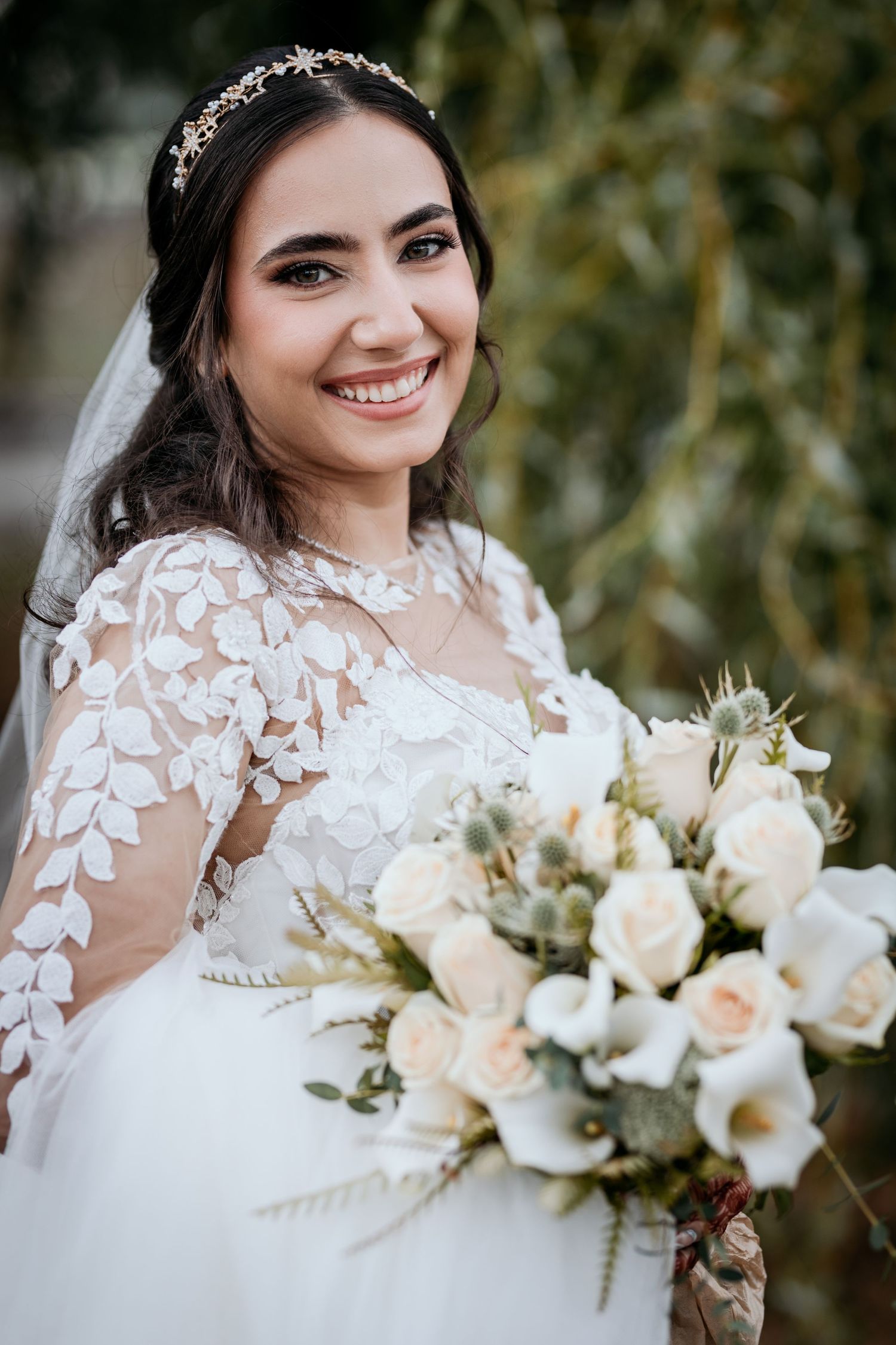 A bride in a white lace gown holds an elegant bouquet of white and blush flowers against a natural outdoor backdrop.