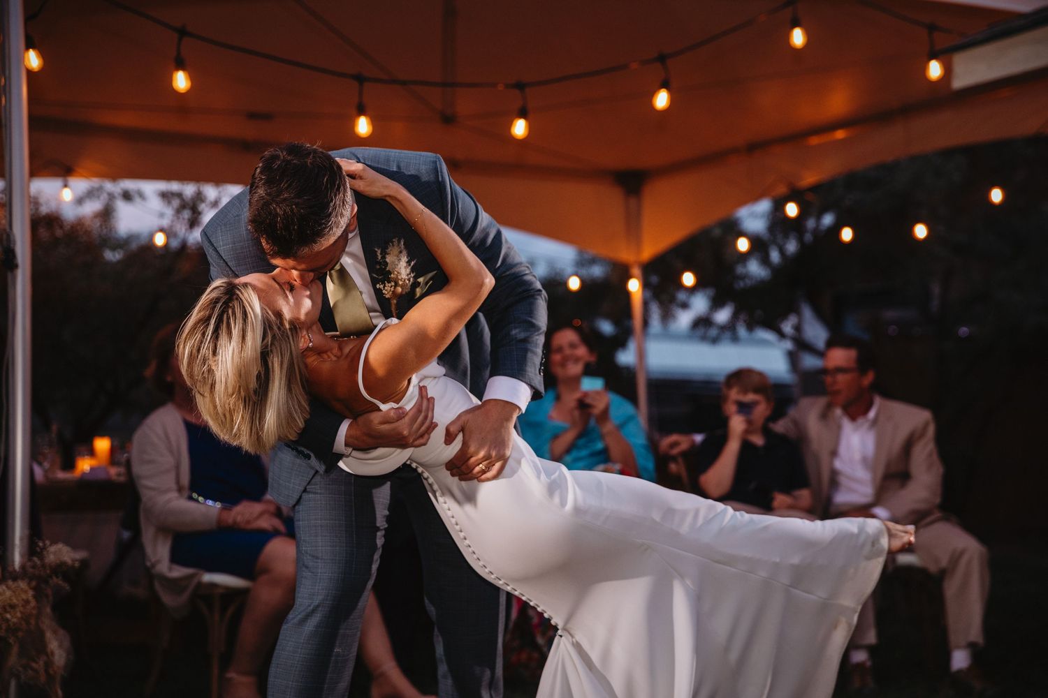 A groom in a blue suit dips his bride during their first dance at their wedding reception.