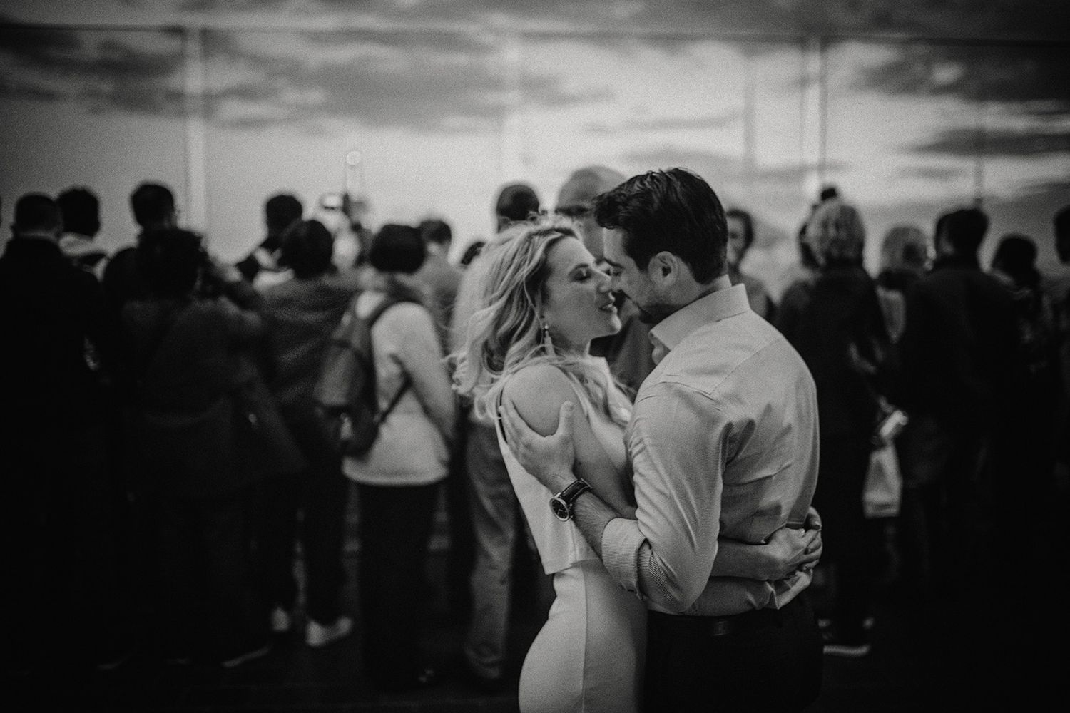 A romantic black and white photo of a couple embracing while a crowd behind them take pictures of the New York skyline.