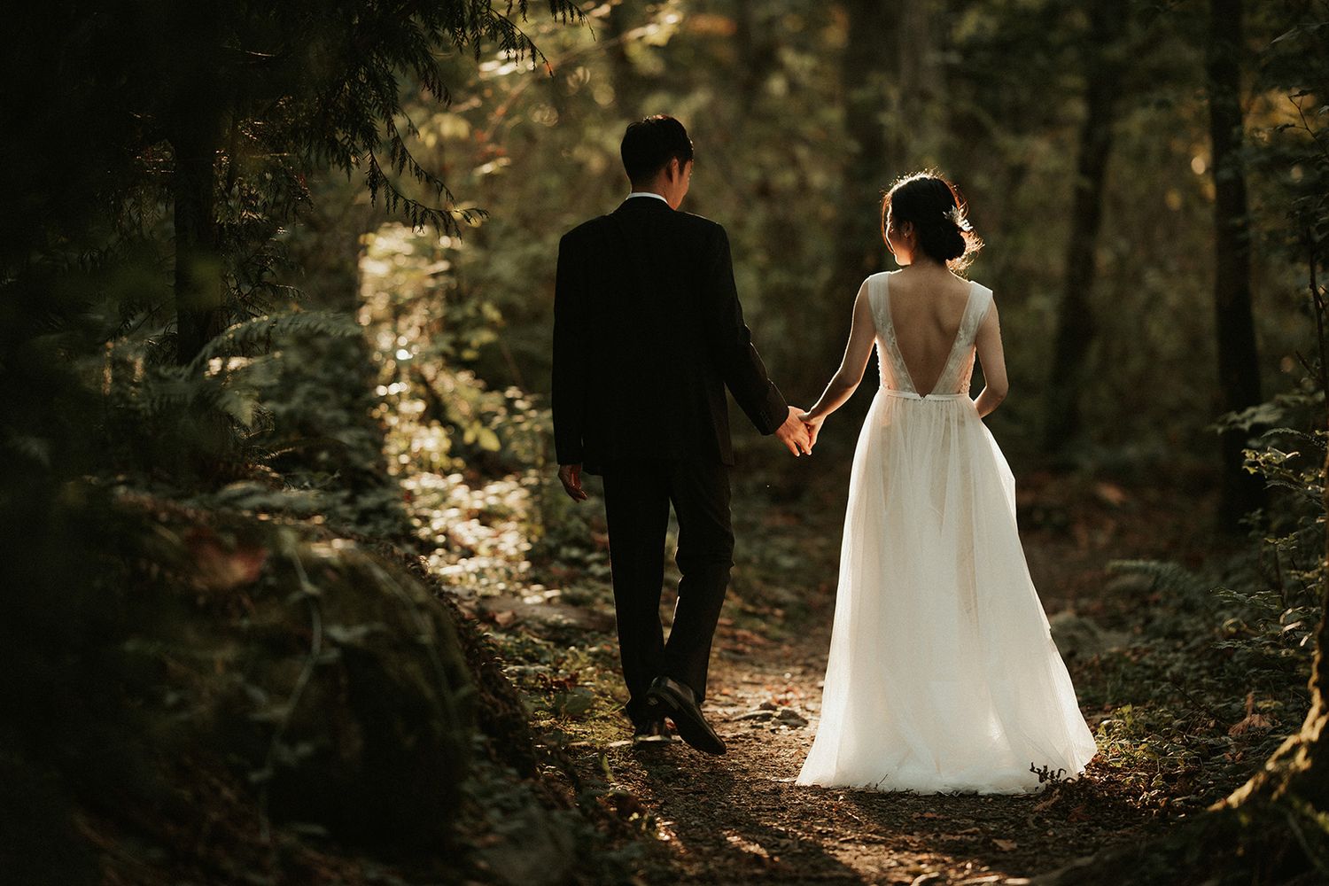 A couple in wedding attire walks through a sunlit forest path holding hands during their intimate outdoor ceremony.