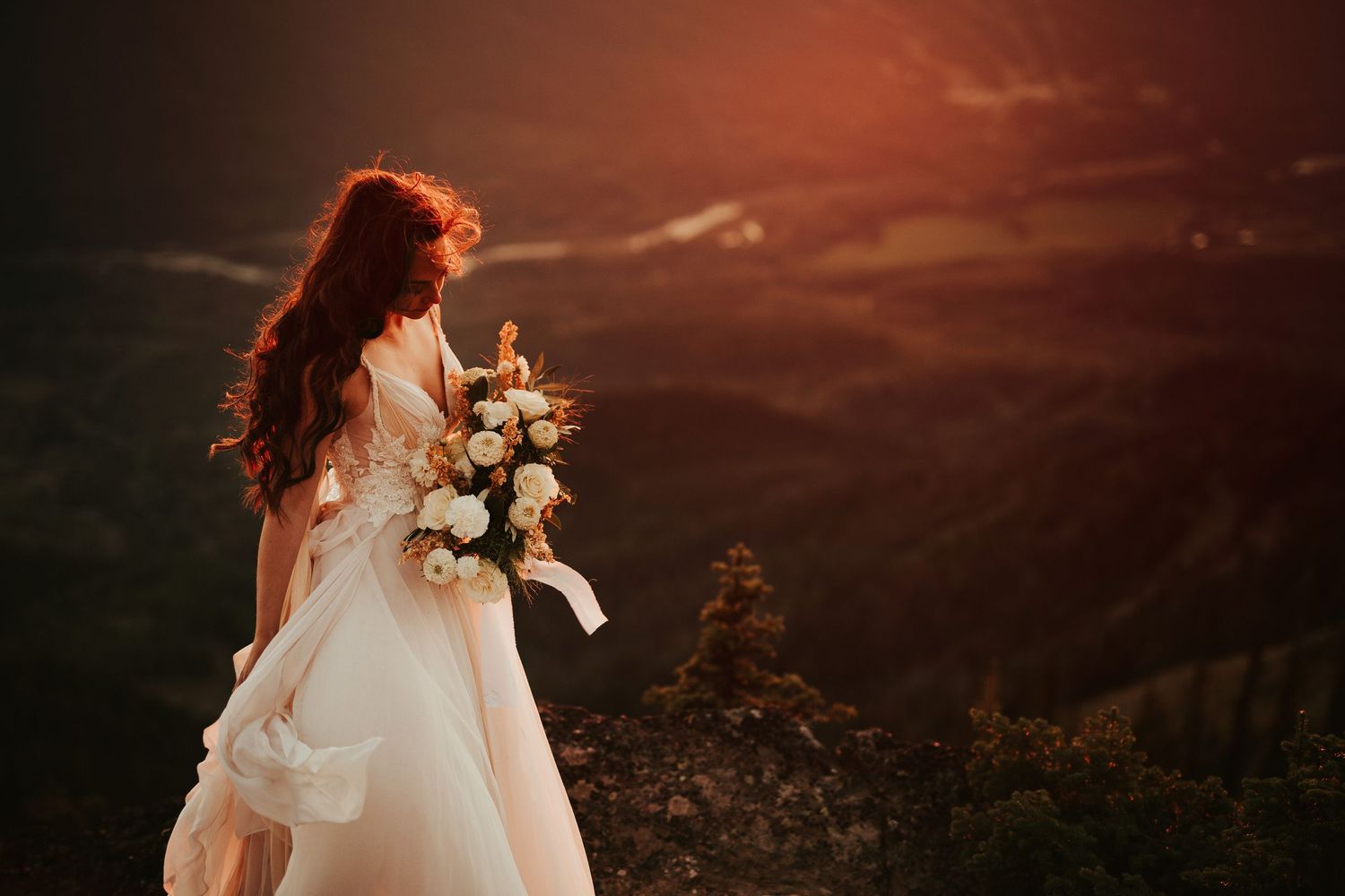 A bride in a flowing white wedding dress stands silhouetted against a dramatic sunset sky in a field.