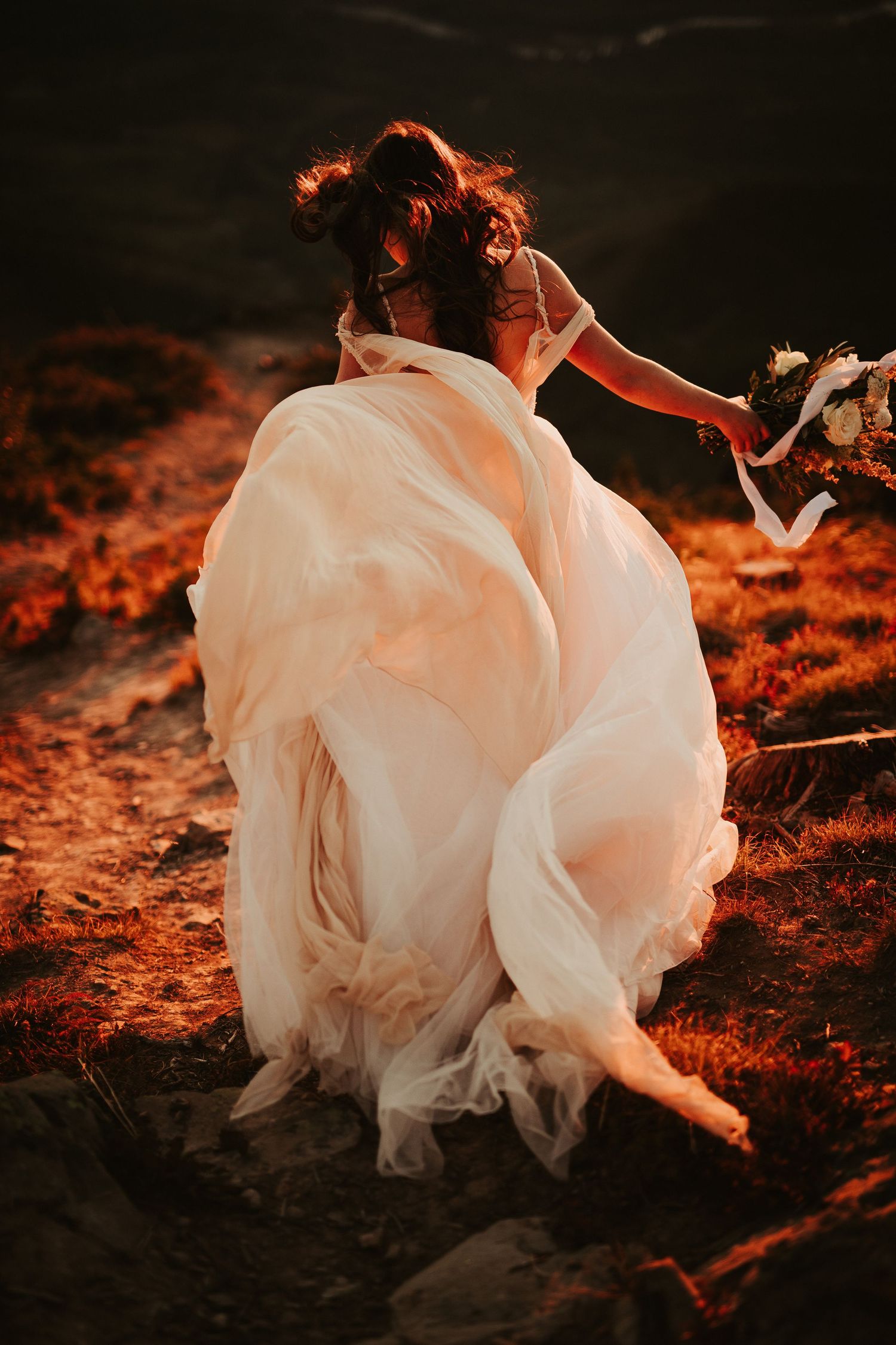 Flowing white wedding dress billowing in the wind against warm sunset lighting on rustic ground.