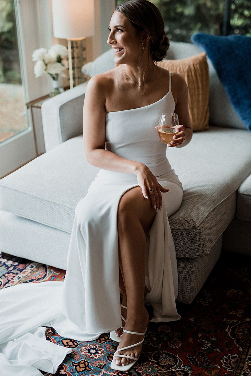 Elegant bride in one-shoulder white gown sits on grey couch holding wine glass and smiling by window.