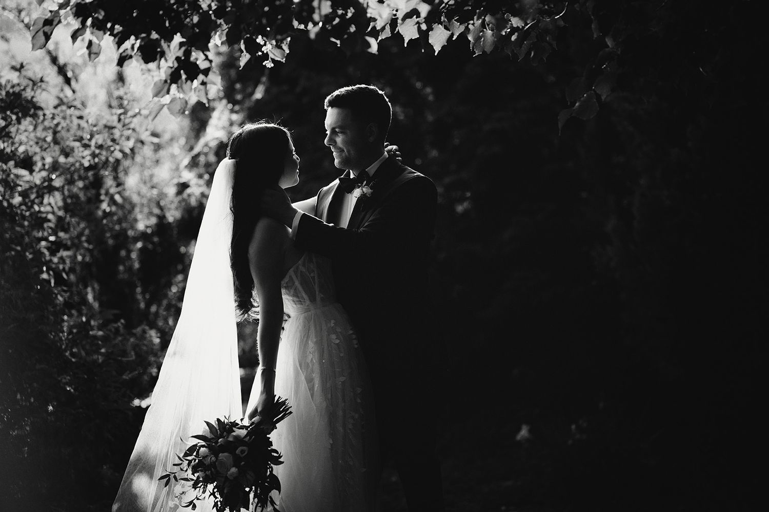 A romantic wedding couple embraces under sunlit trees in a dreamy black and white photograph.