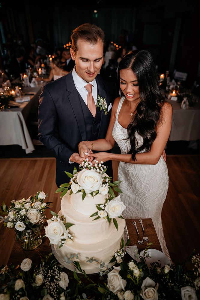 Newlyweds cut their elegant white wedding cake adorned with fresh white roses and greenery at their reception.