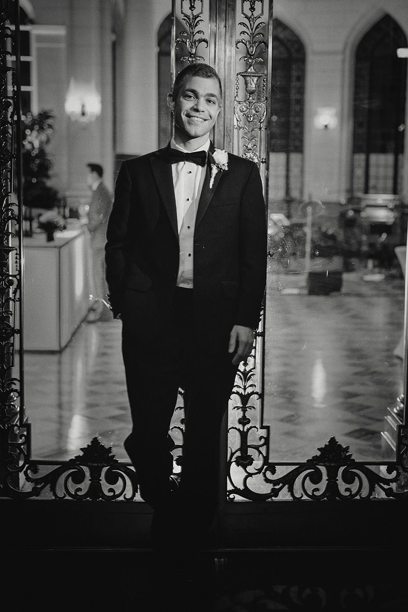 Black and white photo of a man in a tuxedo standing by ornate wrought iron railings in an elegant hallway.