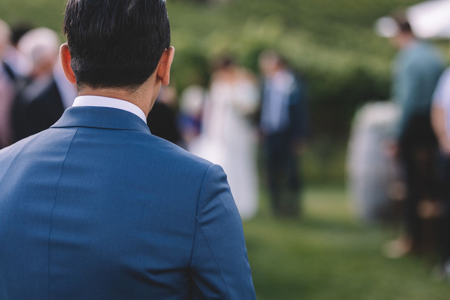A groom in a navy blue suit stands in front of a crowd as he watches his bride approach.