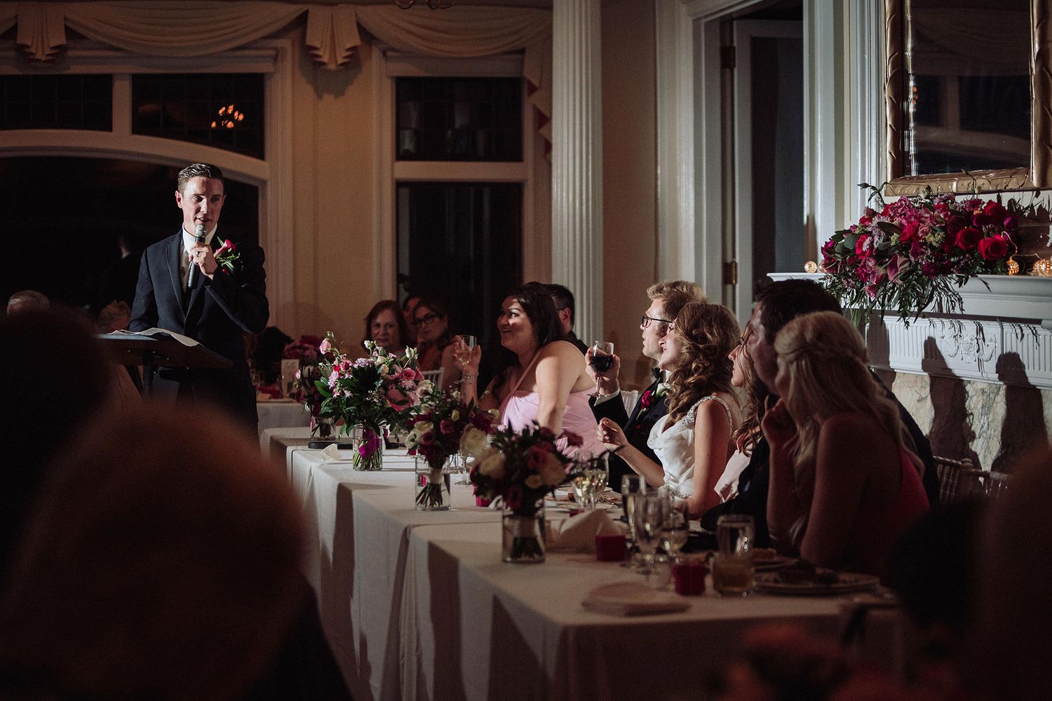Wedding reception guests seated at tables with pink floral centerpieces in an elegant indoor venue. A man gives a speech.