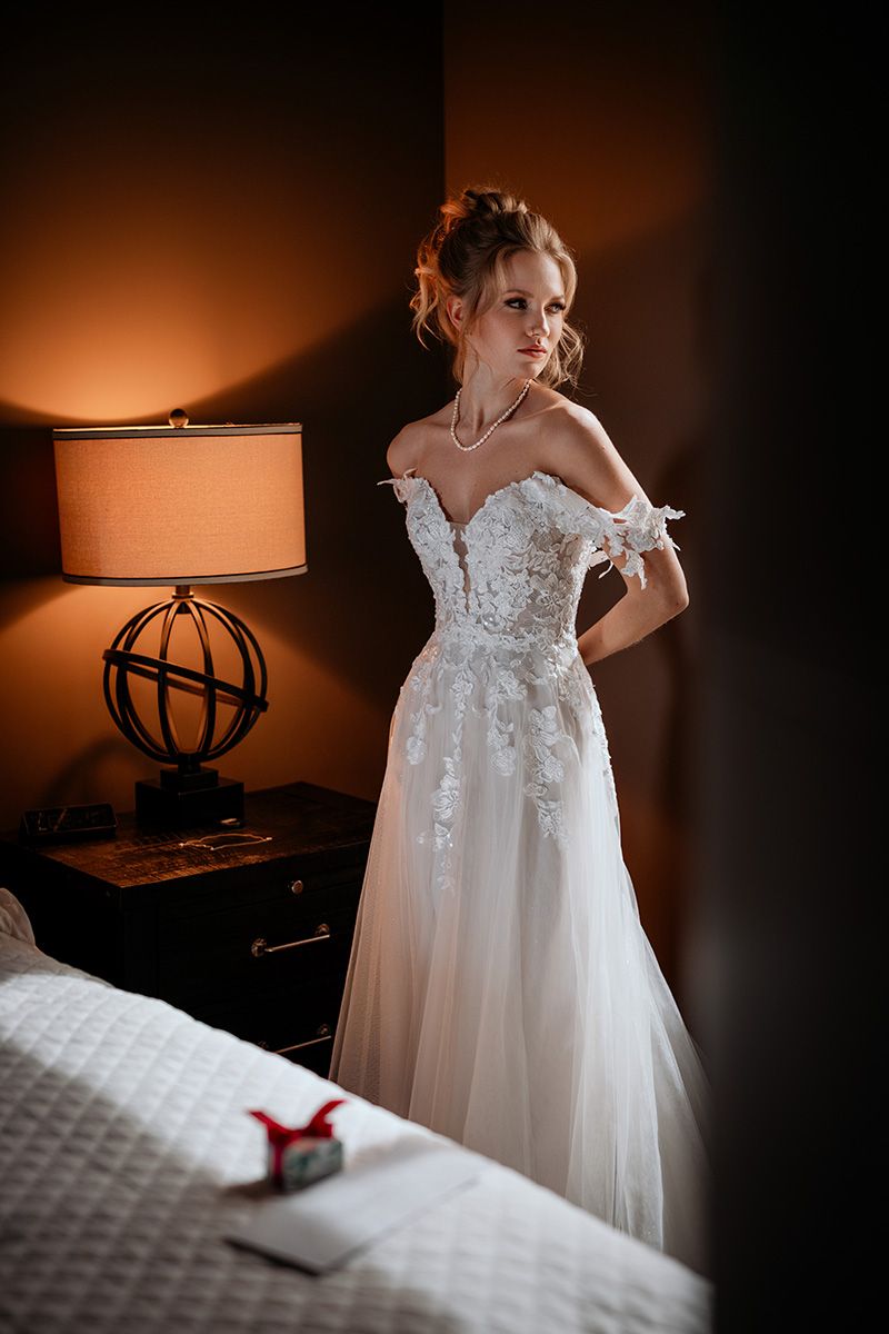 A delicate bride wearing a white wedding dress with lace details and a tulle skirt lit warmly in a room with a bedside lamp.