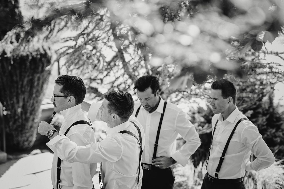 Wedding party in black and white wearing suspenders and bowties laughs together outdoors in a candid moment.