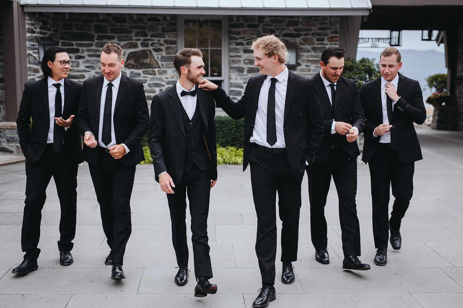 Groomsmen in black suits and ties walking together while laughing and smiling outside a stone building.
