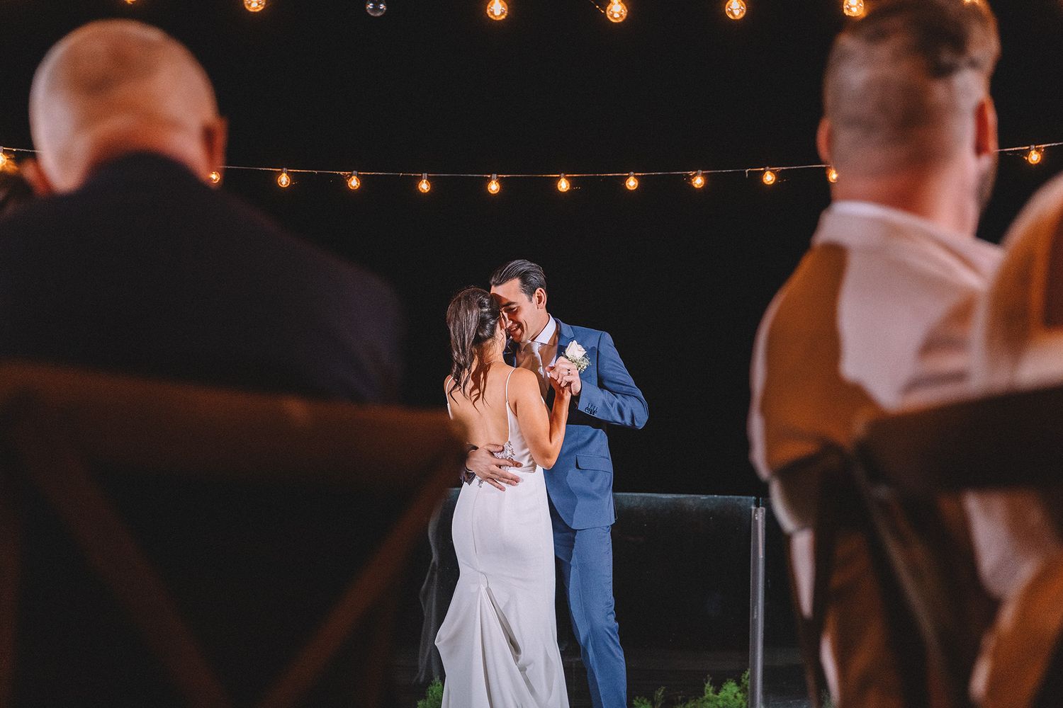 Couple shares romantic first dance at outdoor wedding reception under glowing string lights at night.