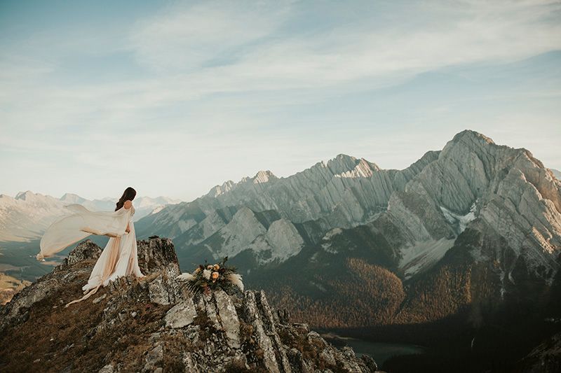 Standing on top of a mountain peak, the wind plays about a bride's wedding gown.