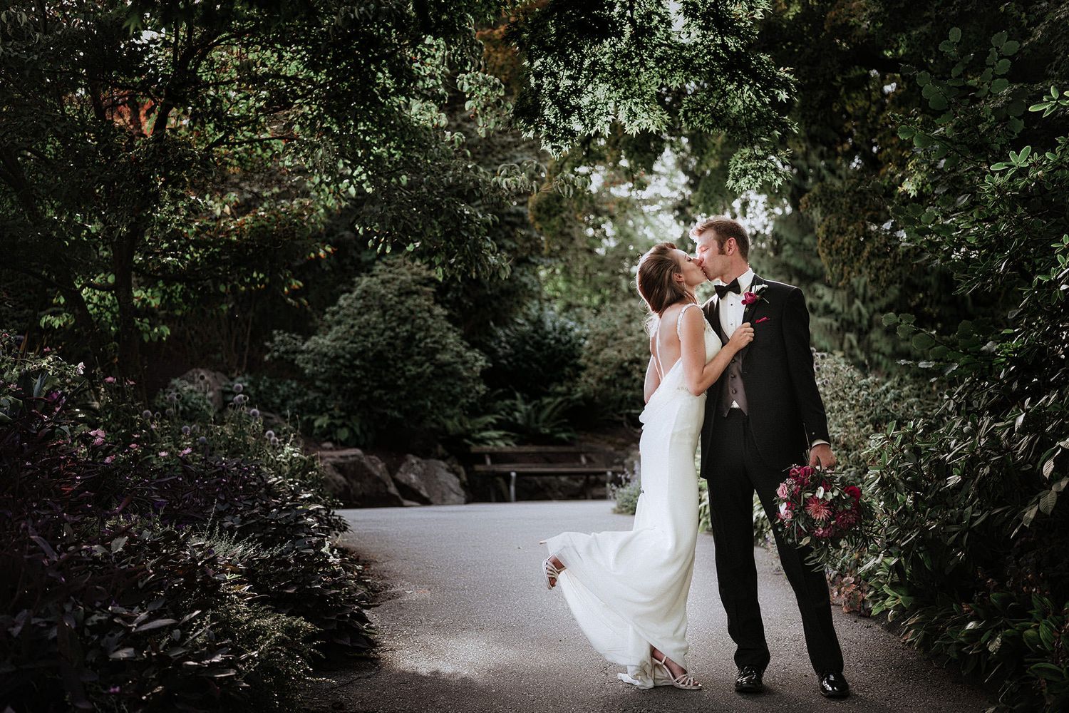 At Queen Elizabeth park a couple walks together on a garden path surrounded by lush greenery and trees on their wedding day.