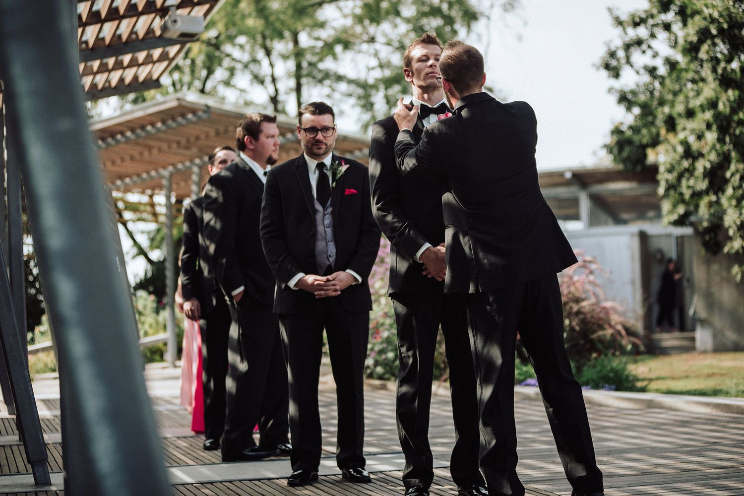 Groomsmen in black suits gather outdoors before a wedding ceremony. The best man adjusts the groom's bow-tie.