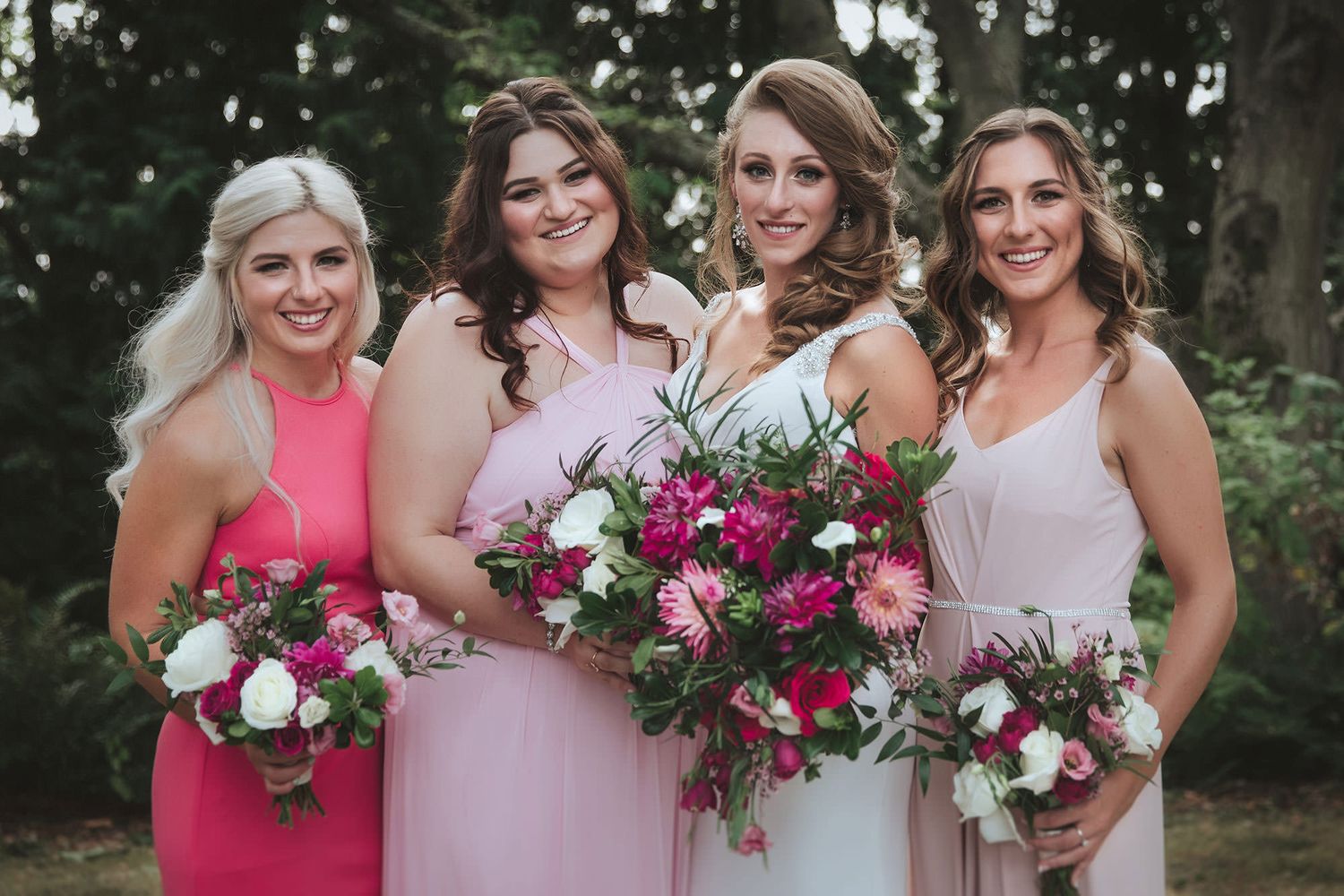 Bridesmaids in pink dresses pose with vibrant burgundy and white floral bouquets during an outdoor wedding celebration.